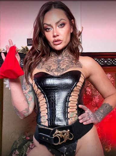 Tattooed woman wearing a black corset with lace-up sides while holding a small red pouch indoors.