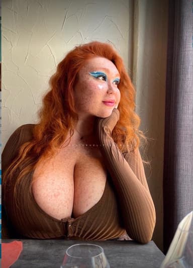 Freckled red-haired woman with blue eye makeup sitting beside a window and looking outside.